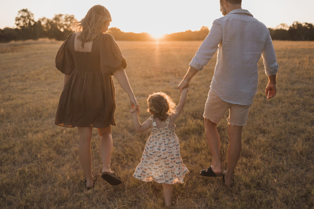 Family photoshoot, with Mum, Dad and child walking through a field at sunset.